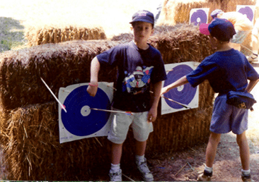 Two boys practicing archery at summer camp with hay bale targets under bright sunlight.