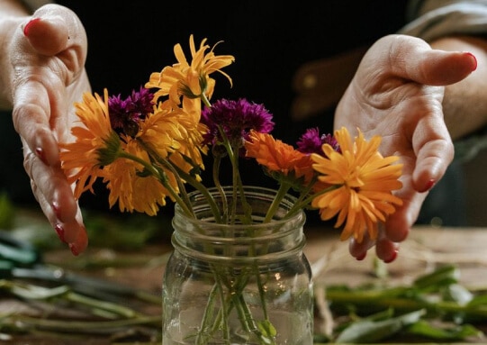 Hands arranging a colorful flower bouquet in a rustic glass jar on a wooden table.