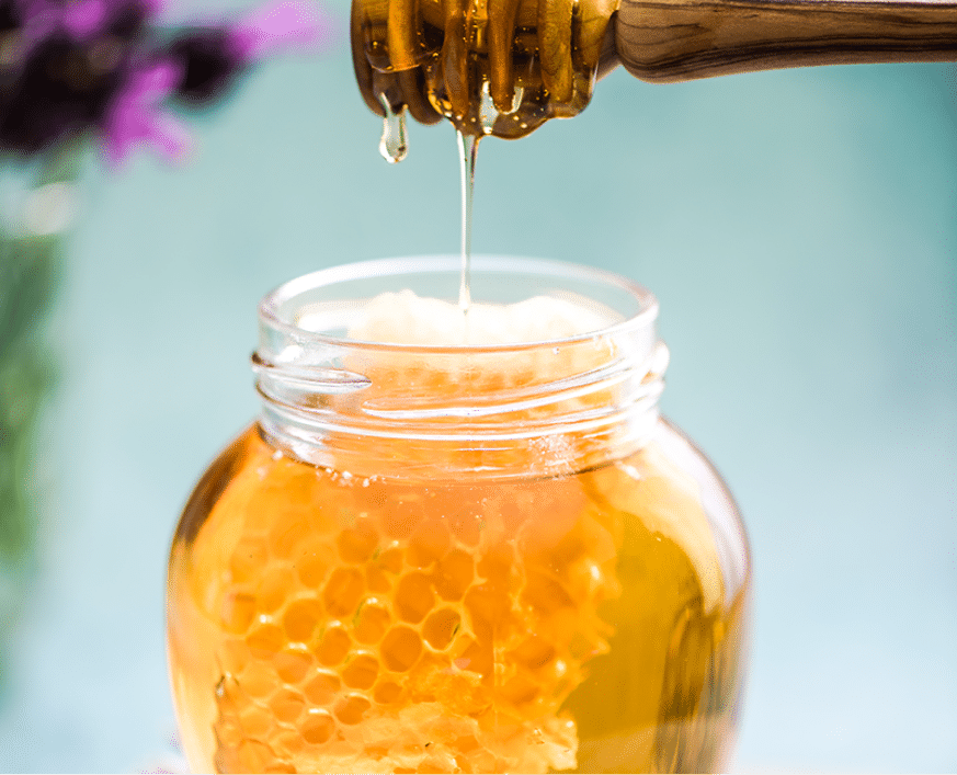 Golden honey dripping from wooden dipper into jar with honeycomb in warm natural light.
