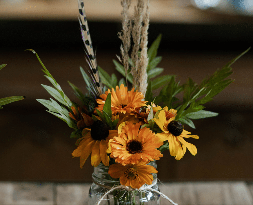 Rustic glass jar with orange and yellow flowers, feather, and twine on a wooden table.