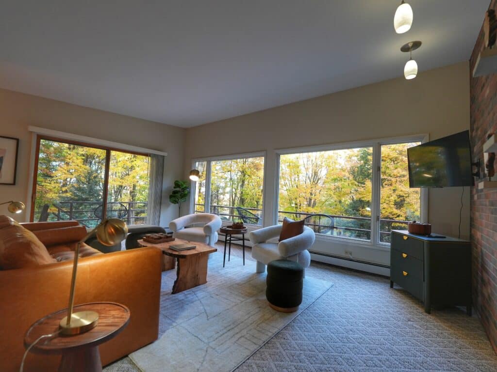 Modern cozy living room with leather sofa, large windows, and scenic autumn forest view.