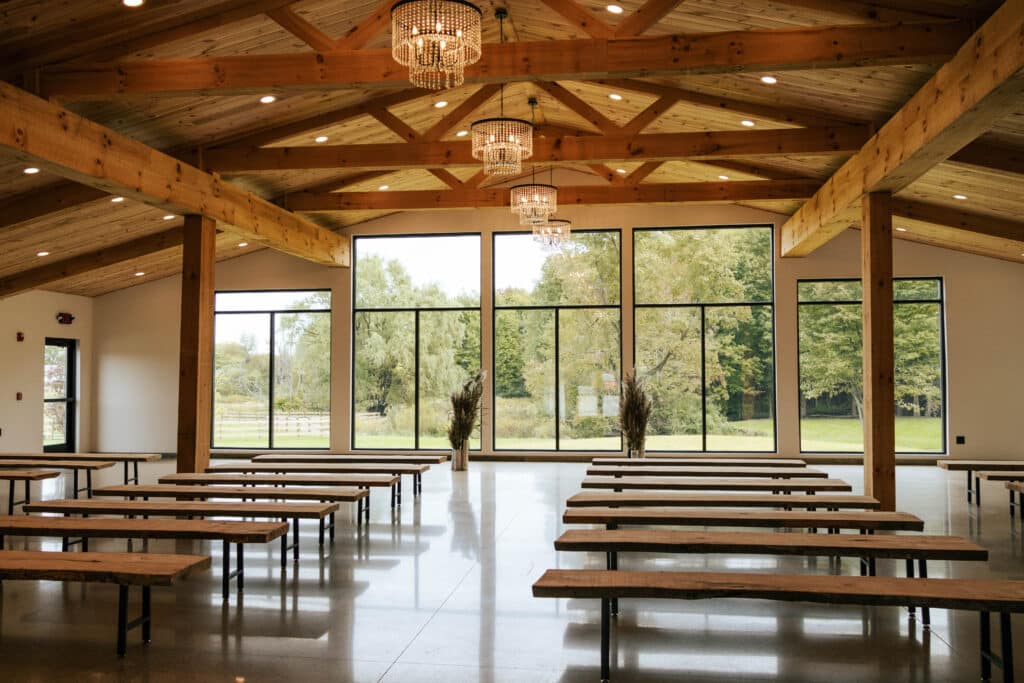 Spacious rustic-modern hall with exposed wooden beams, vaulted ceilings, and large windows overlooking greenery.