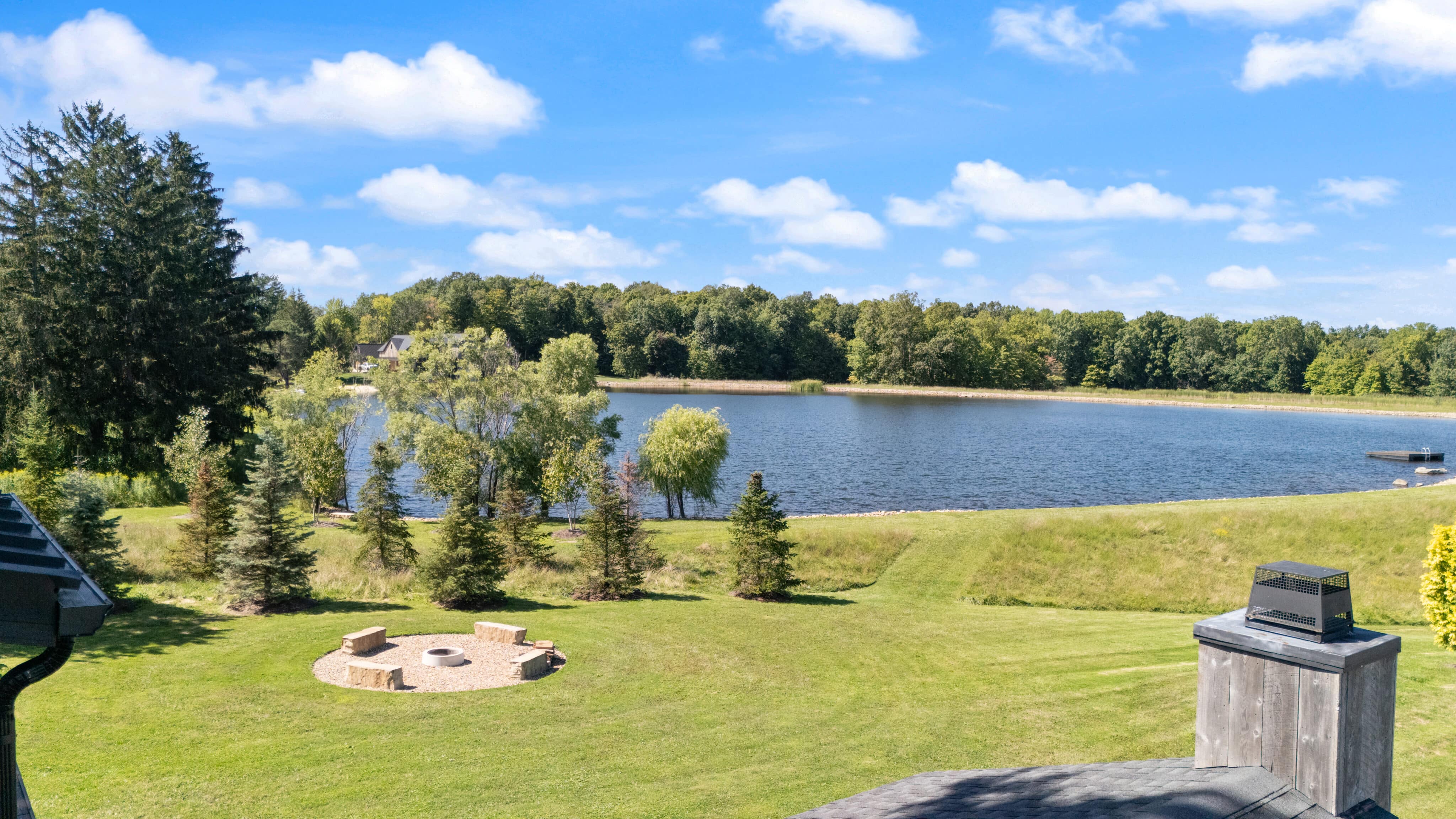 Peaceful lakeside landscape with clear blue water, green grass, trees, and sunny sky.
